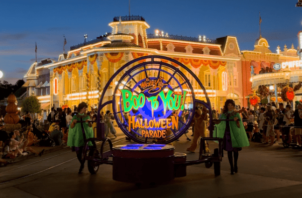 A imagem mostra uma cena noturna do desfile "Boo to You! Halloween Parade" no parque temático da Disney. No centro, há um grande círculo iluminado com luzes roxas e verdes, exibindo o nome do desfile. O fundo revela uma construção clássica do parque, adornada com decorações festivas de Halloween, incluindo abóboras e luzes laranjas. À frente do círculo, duas pessoas estão empurrando o símbolo do desfile, vestidas com roupas temáticas que combinam com o ambiente. O céu escurecido e as luzes ao redor realçam o clima festivo e mágico do evento.